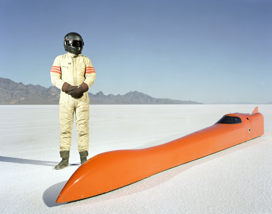 Driver in a race suit beside an orange streamliner on the salt flats